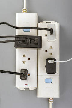 Close-up view of two white power strips mounted on a gray wall, connected together and overloaded with multiple black and white electrical cables plugged into outlets and USB charging ports, showing