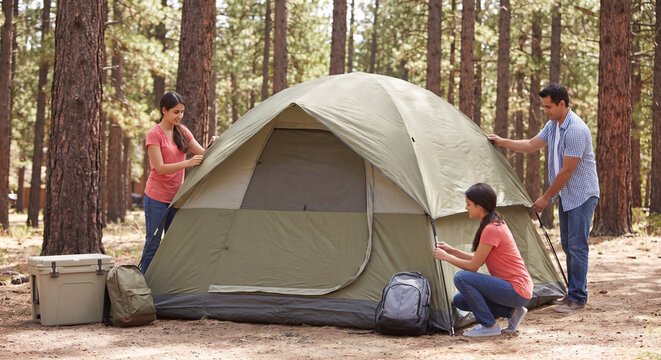 Parents and kids setting up a large camping tent together between tall pine trees at an outdoor wilderness campsite
