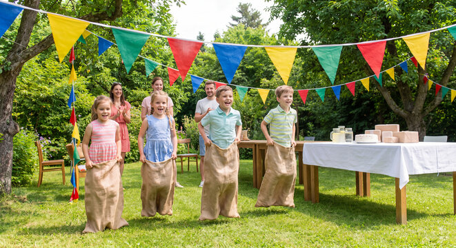 Group of happy children jumping in burlap sacks during potato sack race game on grass lawn at outdoor summer birthday celebration party
