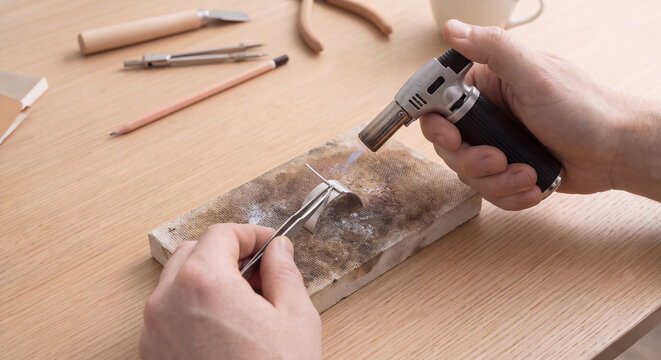 Craftsman goldsmith working with a mini blowtorch soldering precious metal necklace piece in an authentic artisanal jewelry workshop studio