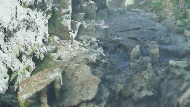 A stream of water flows over rocks in a natural setting. Sunlight shines down on the scene, highlighting the texture of the stones. The area is surrounded by greenery.