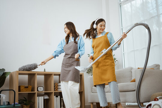 Two women smile as they dust shelves and vacuum the floor at home, blending cleaning, teamwork, friendship and music into a happy lifestyle where listening makes dusting and house cleaning enjoyable.