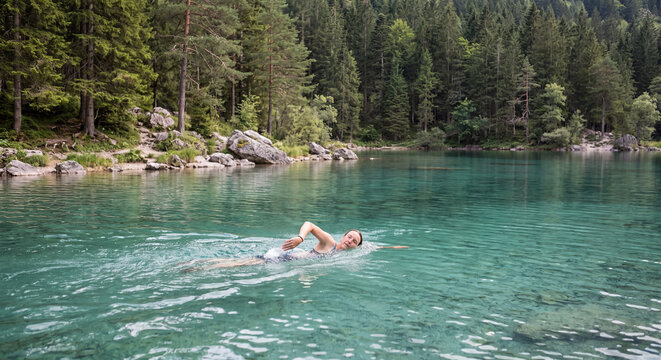 Young woman enjoying a refreshing swim in the turquoise crystal clear waters of a scenic mountain lake surrounded by lush green pine tree forest in summer
