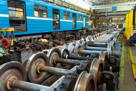 Train wheels and chassis are organized neatly in a railway workshop. Workers move around, busy with repairs and maintenance tasks in the bright space filled with tools and machinery