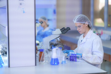 Fototapeta premium Two female medical researchers in blue surgical gowns with hairnets and protective goggles adjusting the scope over blue-capped tubes inside a busy medical diagnostics laboratory.