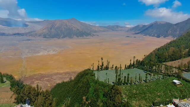 Aerial view of Jembatan Kaca overlooking the vast volcanic caldera of Mount Bromo with lush green hills and clear blue sky in Kecamatan Sukapura, Jawa Timur, Indonesia.