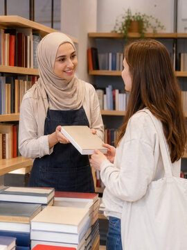 A bookseller wearing a hijab hands a book to a female customer inside a bookstore with shelves and stacked books visible.