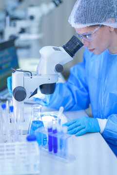 A female research scientist in a white lab coat, hairnet and clear safety goggles bending to the stereo microscope beside a tablet at a brightly lit laboratory workbench.