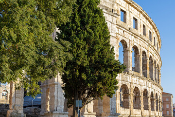 Obraz premium Pula Arena Roman Amphitheater Framed by Green Trees. Pula Arena Roman amphitheater framed by green trees beneath a clear blue sky.