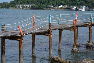 Fototapeta premium Weathered wooden walkway with rope railing extending above rocky coast in Thailand, connecting local waterfront area while overlooking tropical sea. Traditional tropical maritime landscape.