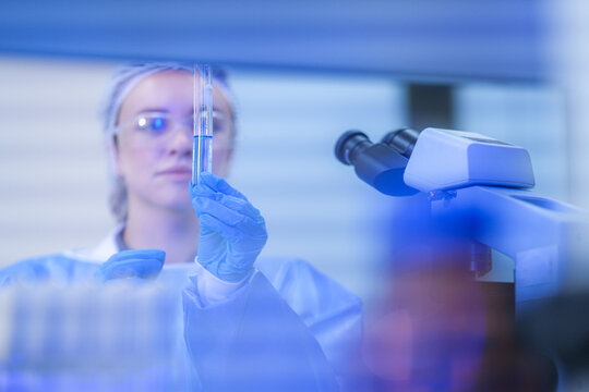 A young woman scientist wearing a white laboratory coat, hairnet and blue nitrile gloves lifting the tube above her eyeline in a modern medical research laboratory.