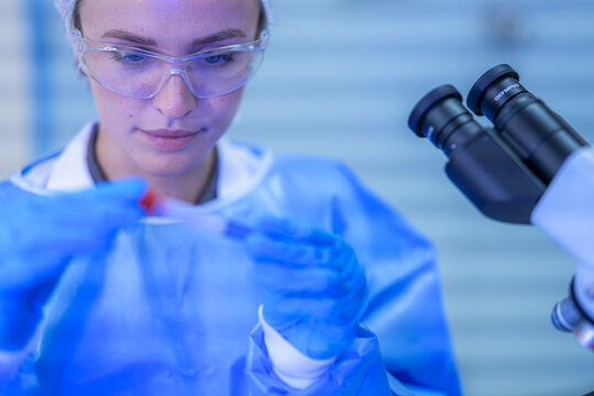 A female medical researcher in a white lab coat, hairnet and clear safety goggles turning the blood tube to read its label at a professional laboratory workstation.