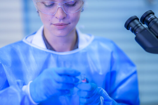 A female medical researcher in a white lab coat, hairnet and clear safety goggles uncapping the red-topped sample tube in a contemporary biomedical research facility.