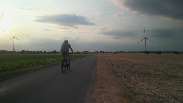 Cyclist Riding Mountain Bike Straight Ahead on Rural Asphalt Road Between Fields Towards Sunset Wind Turbines on Horizon