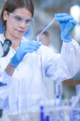 A woman lab technician in white laboratory clothing, safety glasses and blue gloves dispensing into...