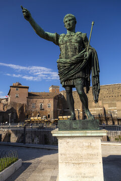 View of the bronze statue of Augustus of Prima Porta standing on a stone pedestal with historic Roman ruins and a clear blue sky in the background in Rome, Lazio, Italy.