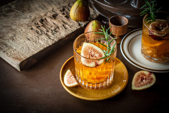 Craft fig cocktail garnished with fresh rosemary sprig and sliced fig in a glass on a dark rustic table. Gourmet bourbon drink with orange peel twist for autumn season menu concept. 