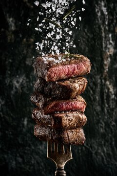 A tall stack of grilled beef steak pieces on a metal fork with coarse sea salt falling from above