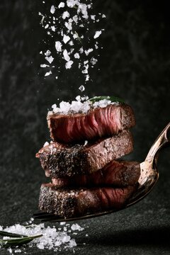 A stack of succulent grilled beef steak pieces being seasoned with coarse sea salt on a dark textured kitchen background