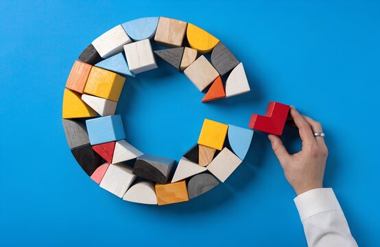 Hand placing red heart shaped wooden block into circular arrangement of colorful cubes cylinders triangles on bright blue background symbolizing completion connection creativity logical thinking artwo