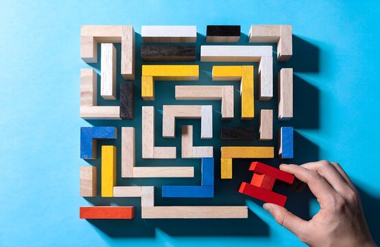Wooden puzzle maze with colorful rectangular blocks on bright blue background hand holding red block symbolizing logical thinking problem solving strategy and spatial reasoning artwork