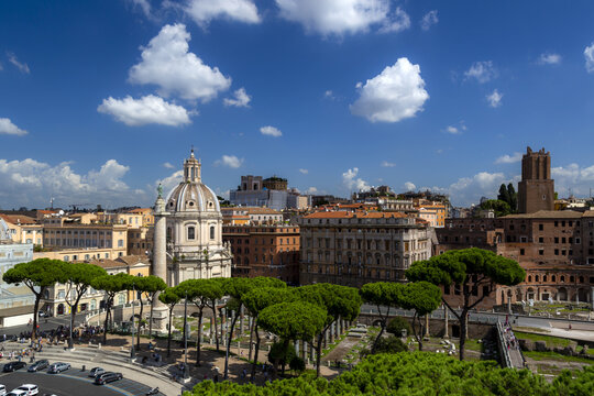 View of Trajan's Column and the Church of the Most Holy Name of Mary at the Trajan Forum, surrounded by historic buildings, pine trees, and blue sky in Rome, Lazio, Italy.