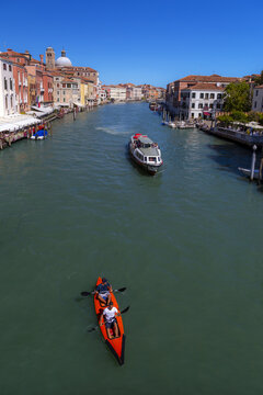 View of two people paddling in a bright orange kayak on the Grand Canal, with a water bus passing by and historic buildings lining the banks in Venice, Veneto, Italy.