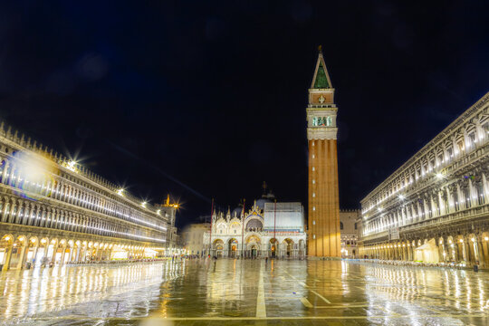 View of the illuminated St. Mark's Square and St. Mark's Campanile at night with reflections on the wet pavement in Venice, Veneto, Italy.