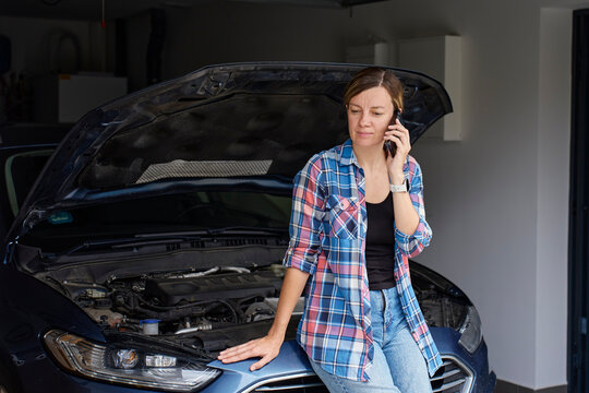 Woman talking on smartphone beside car with open hood in home garage. Car trouble concept