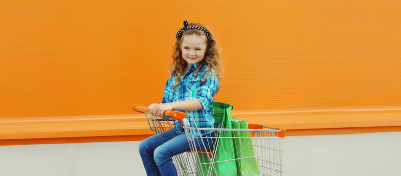 Happy little girl child with trolley cart and colorful shopping bags on city street