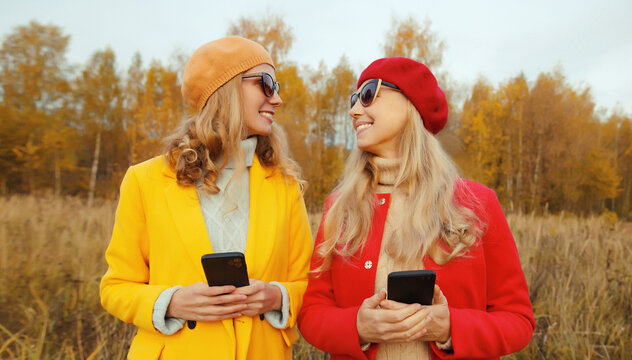 Happy women friends talking with phone in autumn park, stylish girlfriends smiling holds smartphone
