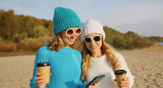 Happy young women friends with smartphone together on beach, smiling girlfriends using phone on sea