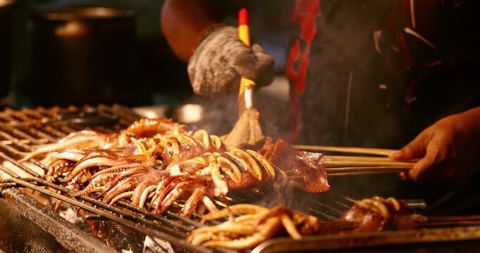 Grilled squid skewers cooking on charcoal grill at night market. Fresh seafood prepared by street vendor. Asian street food culture, culinary travel, outdoor cooking, smoke and fire.