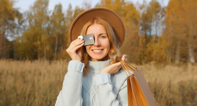 Smiling young woman with credit bank card, shopping bag in autumn park, girl happy with purchases