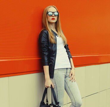 Stylish young woman standing in black leather jacket, sunglasses, modern blonde girl posing in city