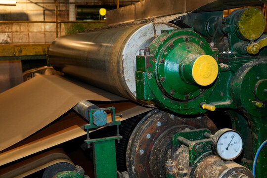 Brown paper moves through massive metal rollers in a factory setting as recycled materials are transformed into durable cardboard packaging sheets
