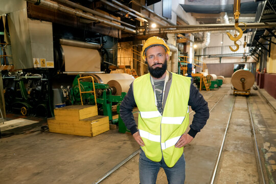 Worker wears a yellow safety vest and hard hat while standing in a busy cardboard recycling plant surrounded by large paper rolls