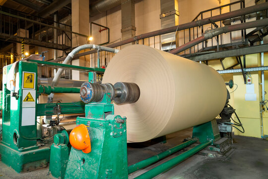 Industrial machine rolls large cardboard sheets inside a recycling plant where workers process paper materials for shipping during a daytime manufacturing shift