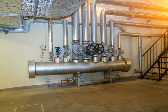 Metal pipelines and hand wheels installed along a light wall inside a basement utility room manage liquid flow for building temperature control