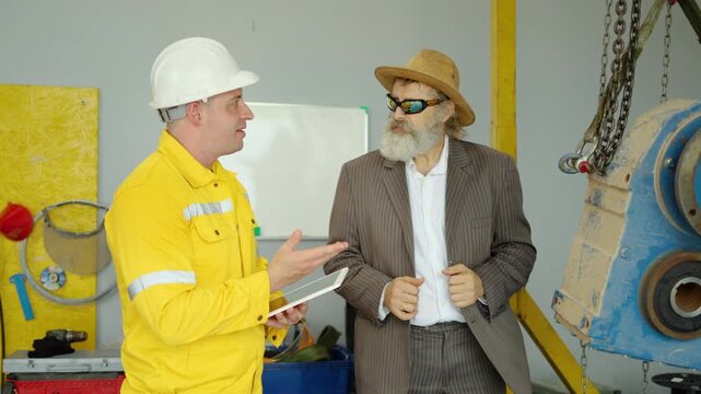 Senior manager and industrial engineer with digital tablet inspect heavy machinery suspended on chain hoist during factory safety audit and manufacturing quality control evaluation in a workshop.
