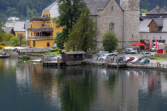 View of the historic stone church and colorful buildings along the shore of Lake Hallstatt with a parking area and calm water in Hallstatt, Upper Austria, Austria.