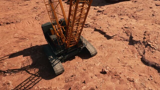 A large crane is positioned on a rocky ground at a construction site. The cranes arm stretches upwards while it prepares to lift materials. Dust surrounds the machinery.