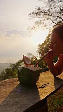 Woman Sips Fresh Coconut Water Through a Straw at a Rustic Outdoor Table During Sunset, Warm Backlight and Tropical Scenery Support Natural Refreshment and Healthy Lifestyle Vertical video.