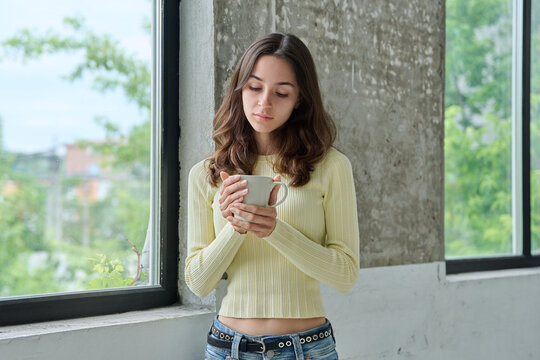 Young thoughtful woman holding coffee mug looking down near window