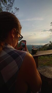 Woman Uses Phone to Photograph Fresh Coconut on Rustic Table During Sunset, Overlooking Forested Hills and Supporting Outdoor Travel Lifestyle Vertical video.