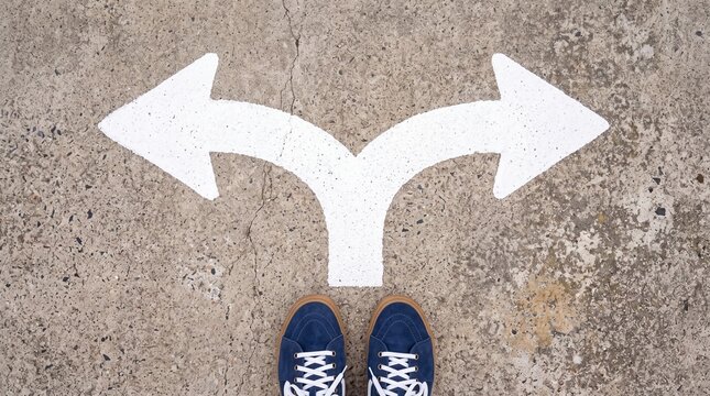 A person wearing blue sneakers stands at a crossroads, looking at white arrows pointing in opposite directions, symbolizing a choice