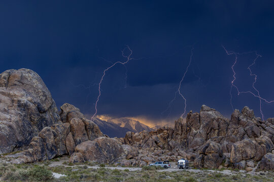 View of lightning strikes over the rock formations of the Alabama Hills with a camper parked nearby under a dark stormy sky in Inyo County, California, United States.
