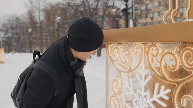 Man in black coat and scarf examines illuminated golden display with white snowflake patterns outdoors during light snowfall. Man touches decorative panel observing its glowing lights
