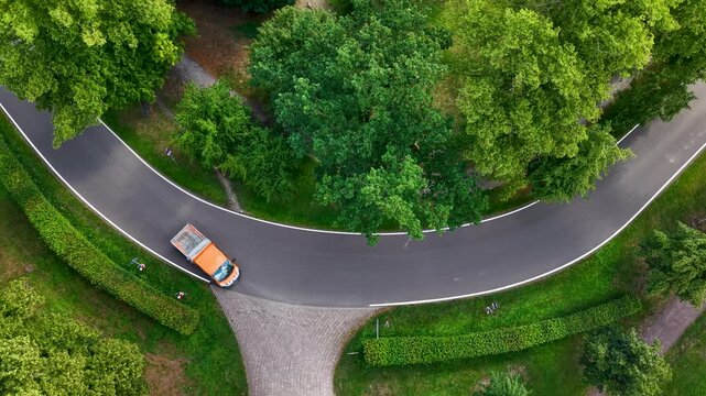 Aerial drone video shows orange utility truck driving along curved asphalt road through dense green parkland. Overhead summer view highlights transport, maintenance, infrastructure, traffic, nature