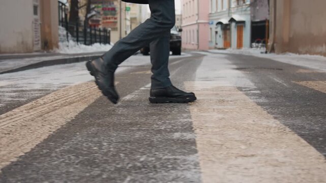 Close up of man legs and feet wearing black leather shoes as man walks across urban street with snow patches on ground. focus remains solely on foot movement capturing each step in detail